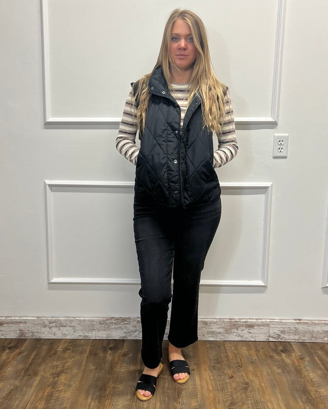 A woman stands indoors wearing a black quilted sleeveless puffer vest over a beige and black striped long-sleeve top, black pants, and black slide sandals, in front of a white paneled wall.