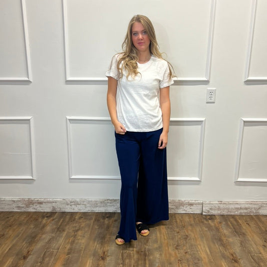 A woman stands in a room with white paneled walls and a wood floor, wearing a white short-sleeve t-shirt and dark navy wide-leg pants.