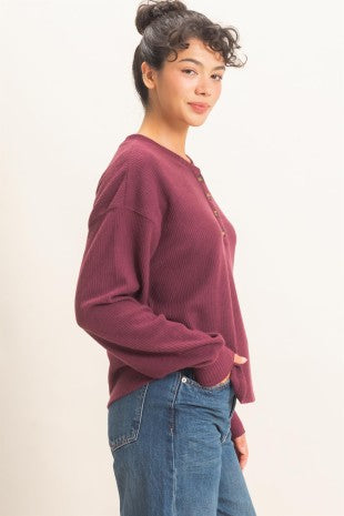 A woman wearing a ribbed knit long-sleeve sweater in burgundy with a buttoned henley neckline, paired with blue jeans, photographed against a plain off-white studio background.