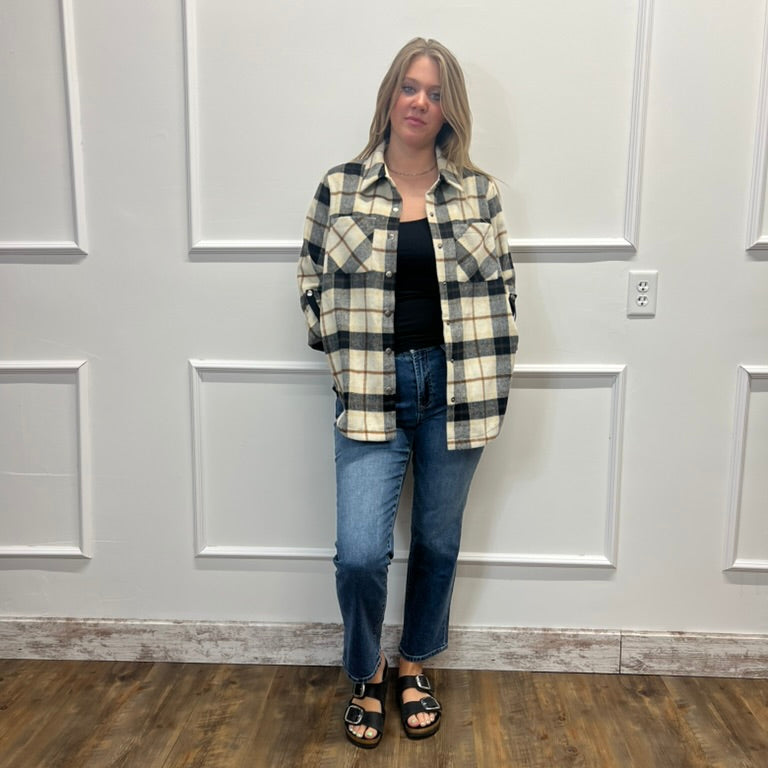 A woman stands against a white paneled wall wearing a black, cream, and gray plaid button-up shirt over a black top, paired with blue jeans and black sandals.