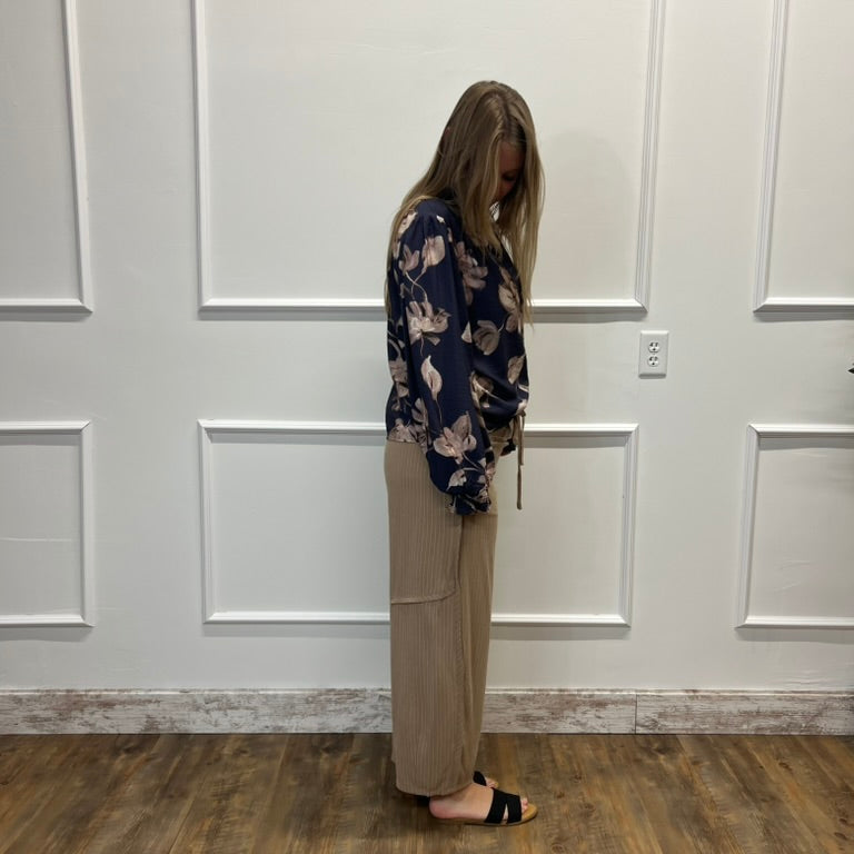 A woman stands in profile against a white paneled wall on a wood floor, wearing a navy floral long-sleeve top and beige wide-leg pants.