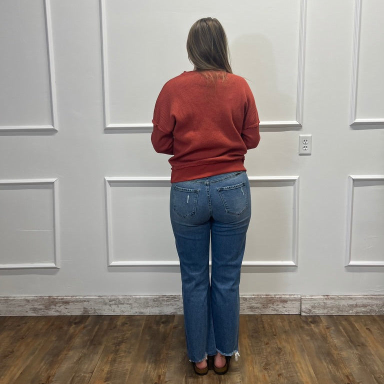 A woman viewed from behind wearing a rust-colored long-sleeve sweater and blue jeans, standing in front of a white paneled wall on a wood floor.