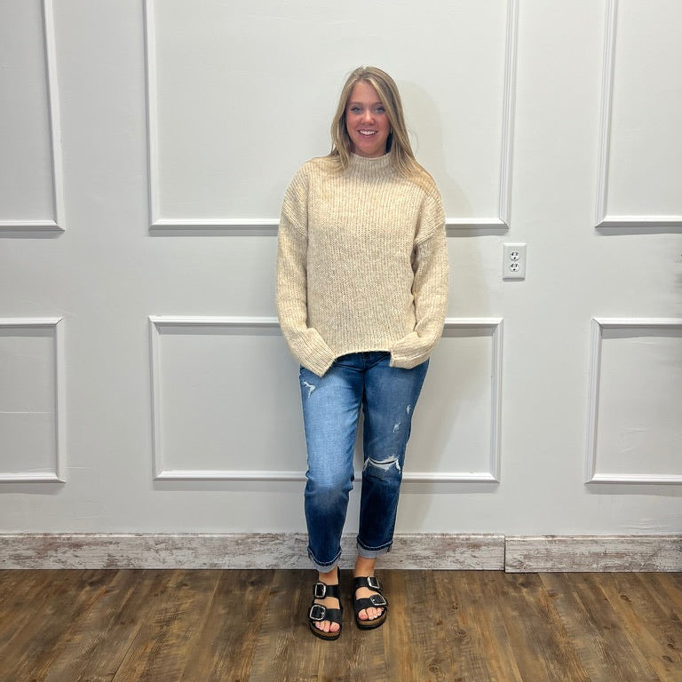 A woman stands in front of a white paneled wall wearing a cream-colored knit mock neck sweater, blue ripped jeans, and black sandals.