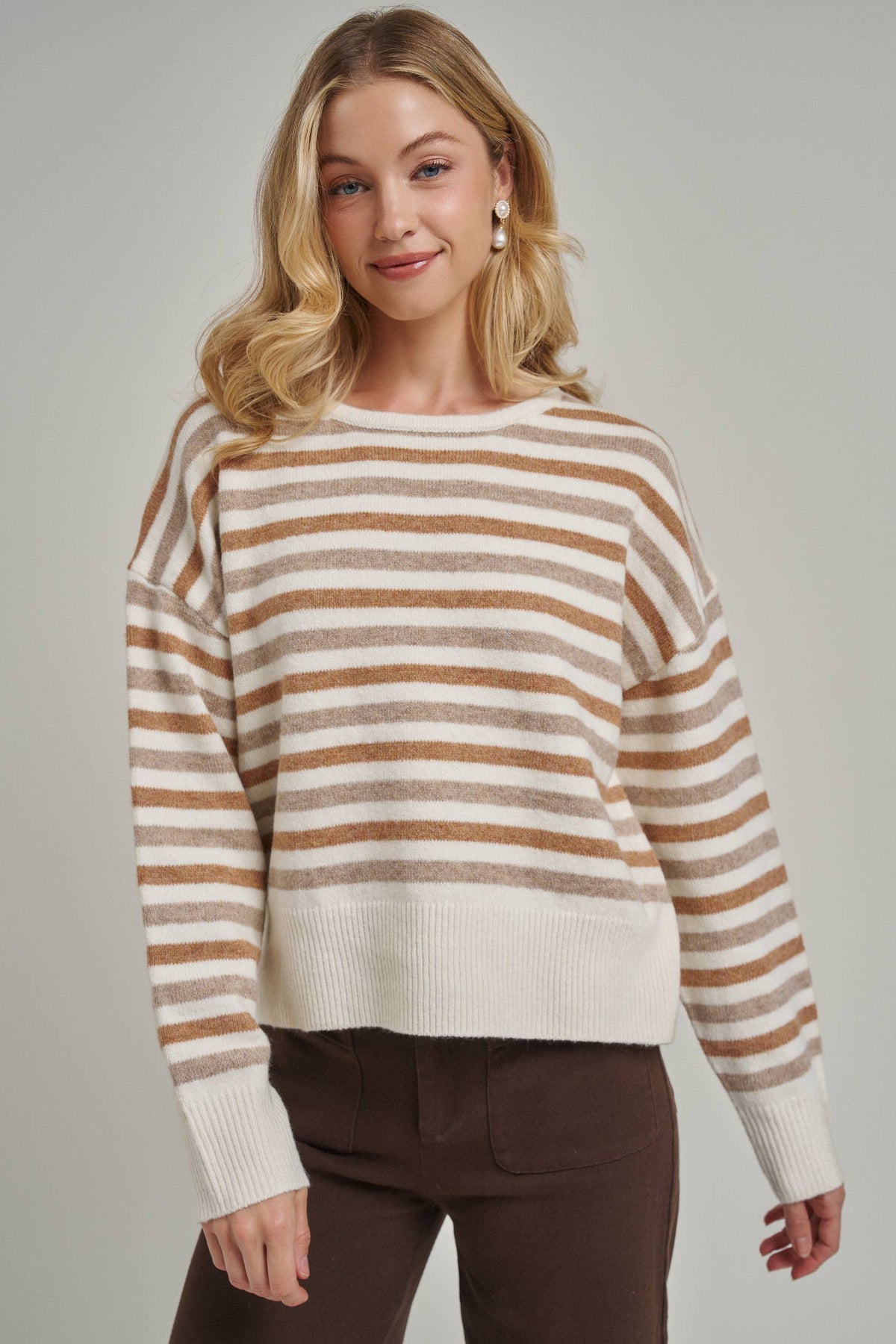 A woman wearing a cream and tan horizontally striped knit sweater with a crew neck and dropped shoulders, paired with dark brown pants, against a solid light gray studio background.