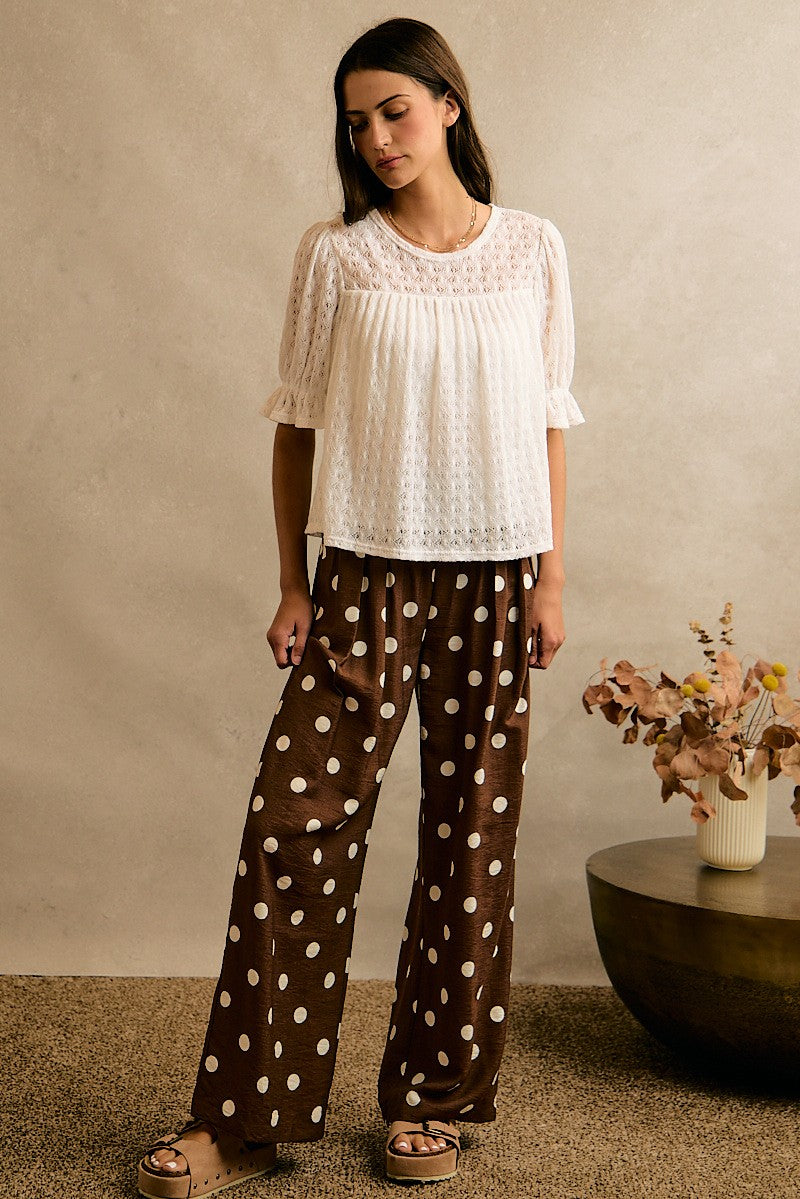 A woman modeling a two-piece lounge set featuring a white short-sleeve lace top and brown wide-leg pants with white polka dots, standing in a studio setting with a textured beige wall and a side table with dried flowers.