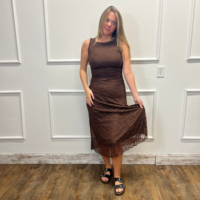 A woman modeling a sleeveless brown lace midi dress with a ruched bodice and sheer straps, standing on a wood floor against a white paneled wall.
