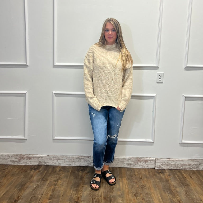 A woman stands in front of a white paneled wall wearing a cream-colored knit mock neck sweater, blue distressed jeans, and black slide sandals.
