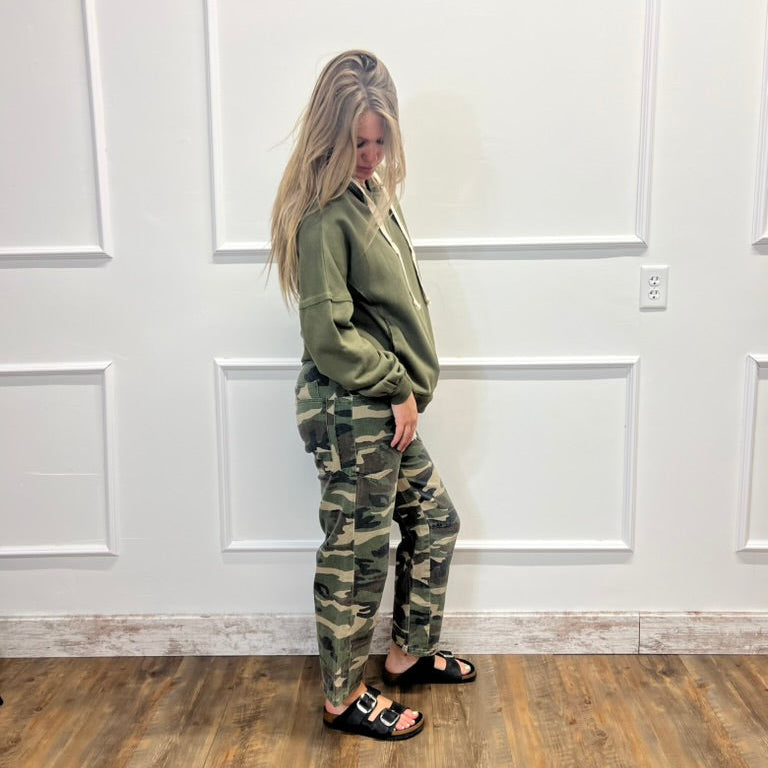 A woman stands in profile wearing an olive green hoodie and matching camouflage cargo pants, paired with black slide sandals, against a white paneled wall with a wood floor.