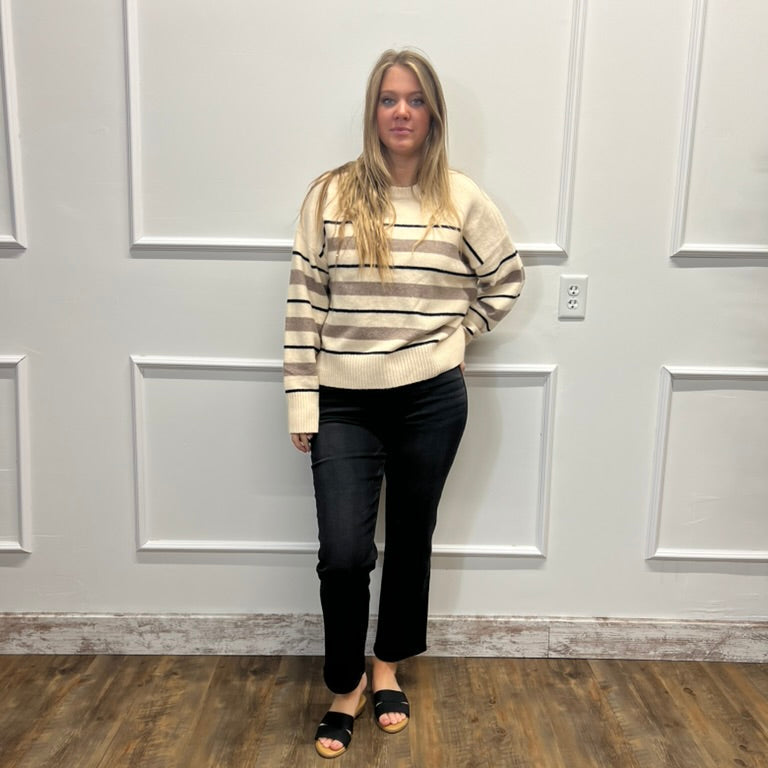 A woman stands in a room with white paneled walls and a wood floor, wearing a cream and brown striped long-sleeve sweater, black pants, and black slide sandals.