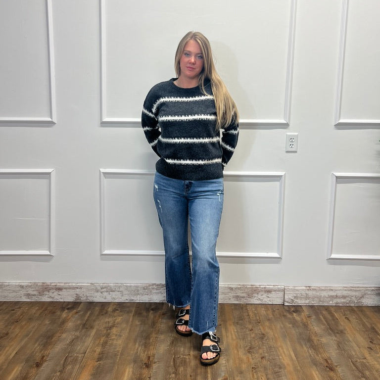 A woman stands in a room with white paneled walls and a wood floor, wearing a black and white patterned knit sweater, blue jeans, and black sandals.