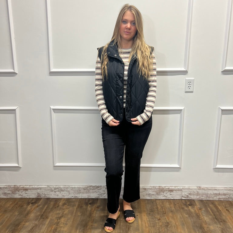 A woman stands indoors against a white paneled wall on a wood floor, wearing a black quilted sleeveless puffer vest over a beige and black striped long-sleeve top, black pants, and black slide sandals.