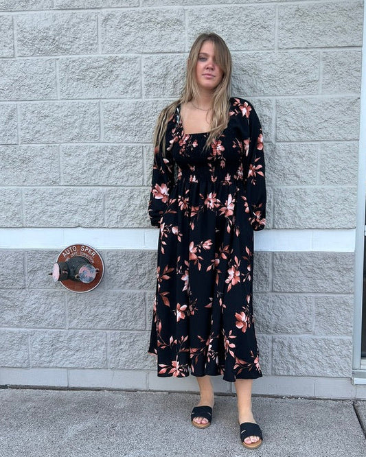 A woman stands outdoors against a light gray stone wall wearing a long-sleeve black midi dress with a pink floral pattern and black slide sandals.