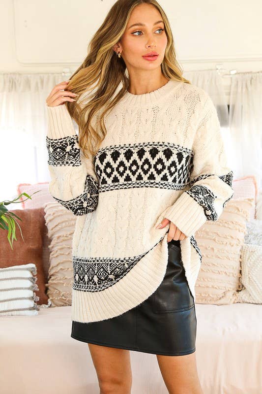 A woman wearing a cream knit sweater with black Nordic geometric patterns across the chest, sleeves, and hem, paired with a black faux-leather mini skirt, standing in a cozy bedroom setting.
