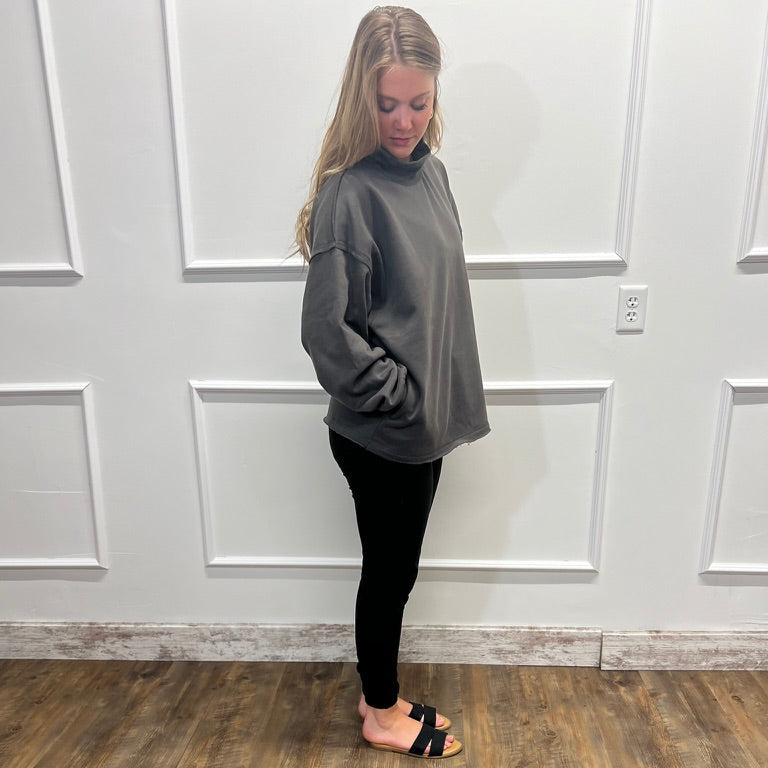 A woman stands in profile wearing an oversized gray mock-neck sweatshirt, black leggings, and black slide sandals, standing on a wood-look floor against a white paneled wall.