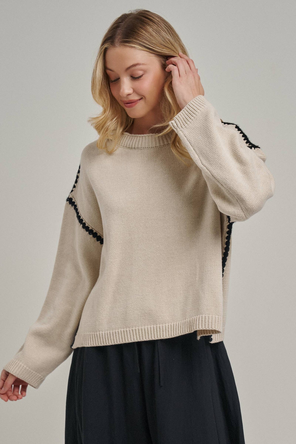 A woman wearing a cream-colored knit sweater with black decorative trim along the shoulders and sleeves, paired with dark navy pants, against a solid light gray studio background.