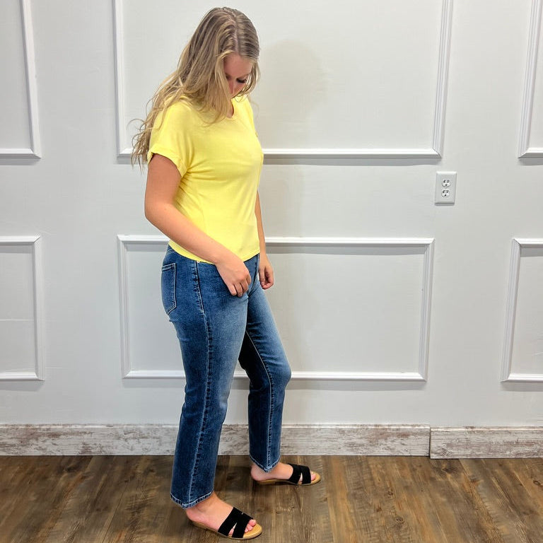 A woman wearing a bright yellow short-sleeve t-shirt and blue jeans stands in a room with white paneled walls and a wood floor, looking down.