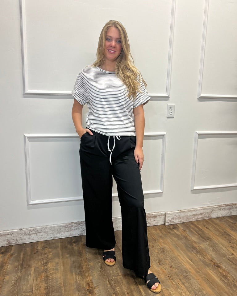 A woman modeling a short-sleeve crewneck t-shirt with thin white horizontal stripes on a light gray background, paired with black wide-leg drawstring pants and black slide sandals, standing against a white paneled wall.
