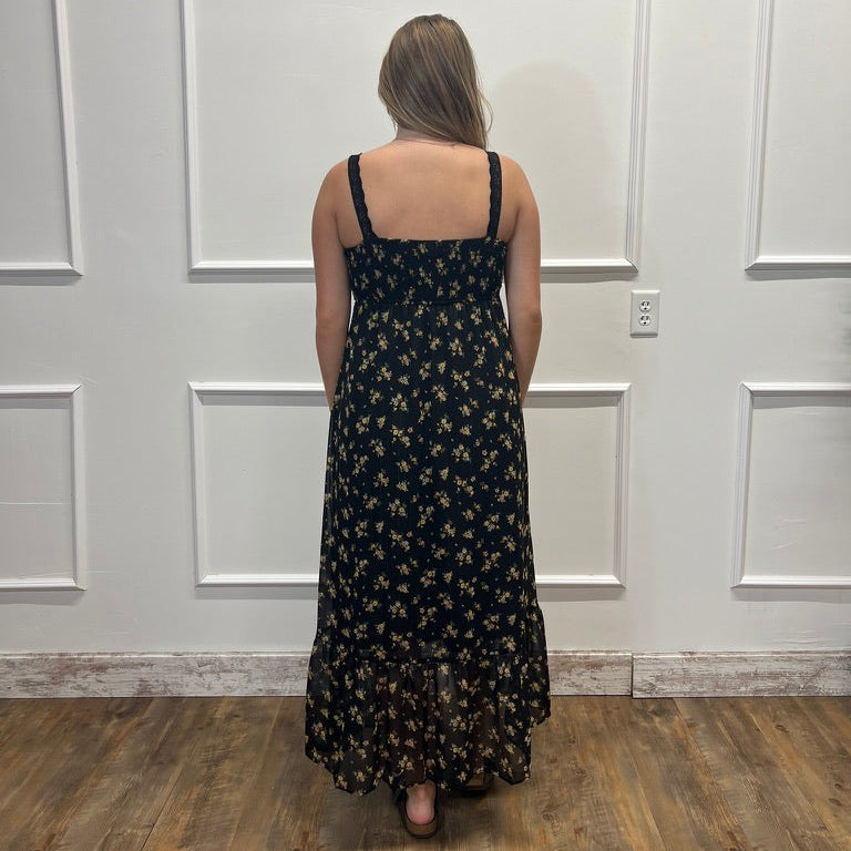 A woman viewed from behind wearing a black spaghetti-strap maxi dress with a small beige floral pattern, standing on a wood floor against a white paneled wall.