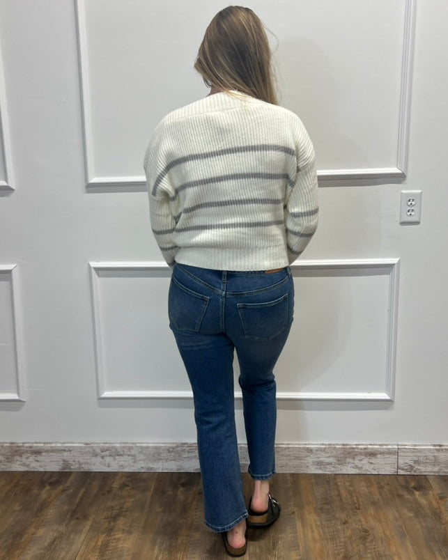 A woman viewed from behind wearing a cream knit sweater with thin gray horizontal stripes and blue jeans, standing on a wood floor against a white paneled wall.