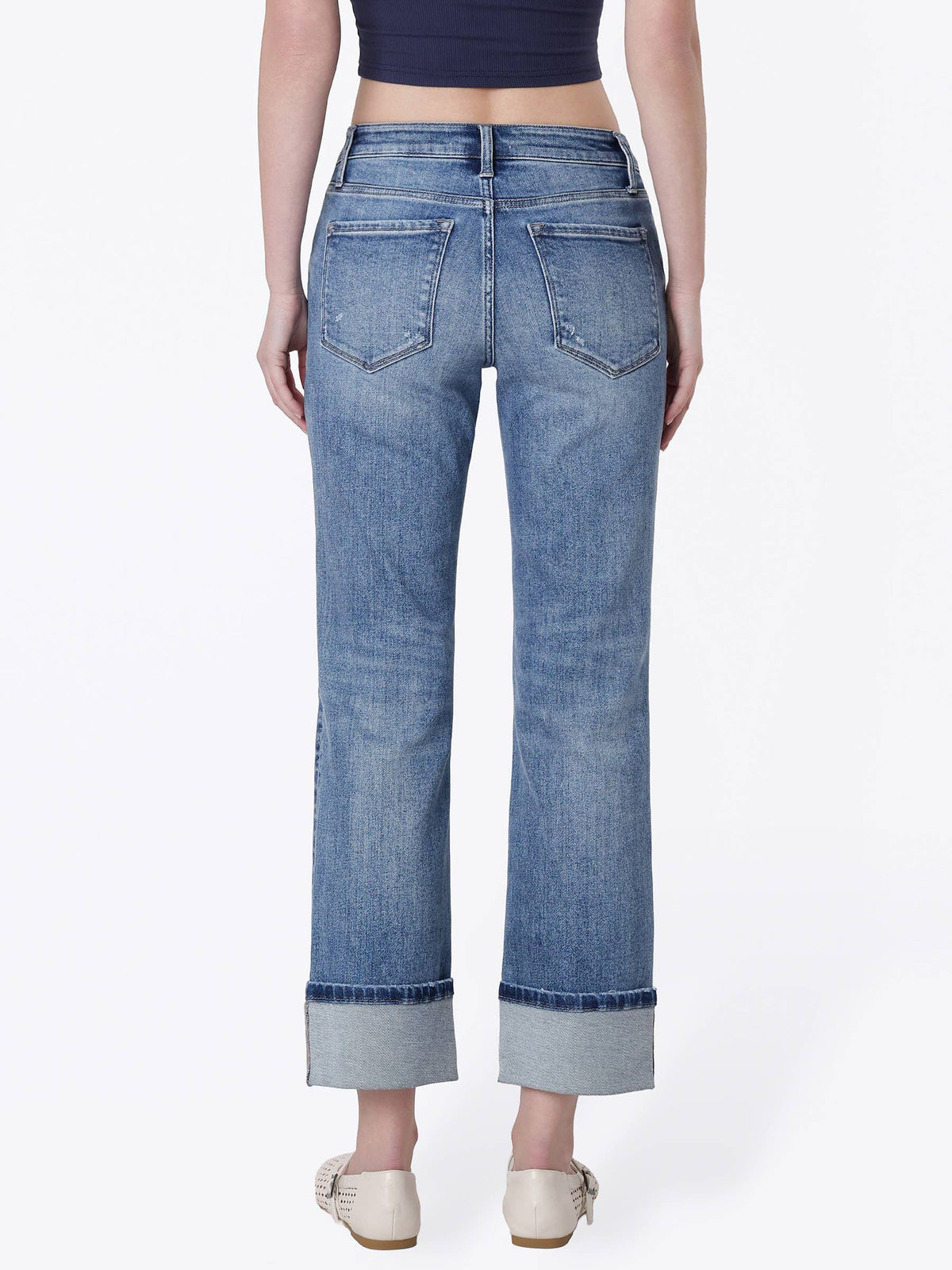 A model shown from the back wearing a pair of high-waisted, wide-leg cropped jeans in medium blue denim with rolled cuffs, paired with a dark navy crop top and beige flat shoes, against a plain white studio background.