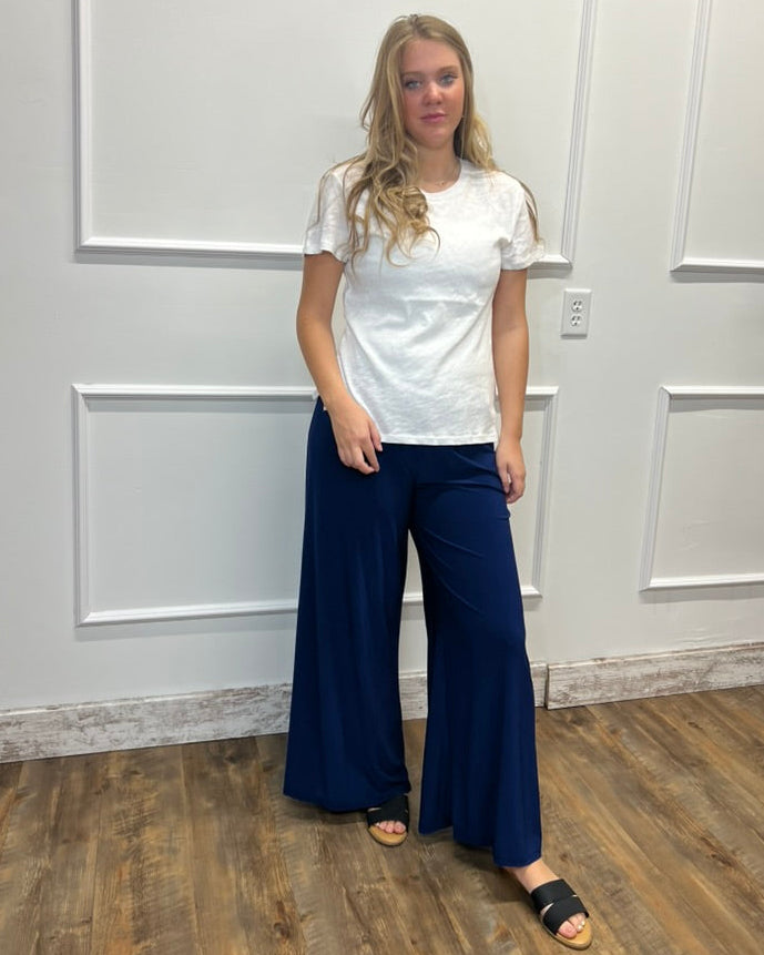 A woman modeling a white short-sleeve t-shirt and navy blue wide-leg pants, standing in front of a white paneled wall with a wood floor.