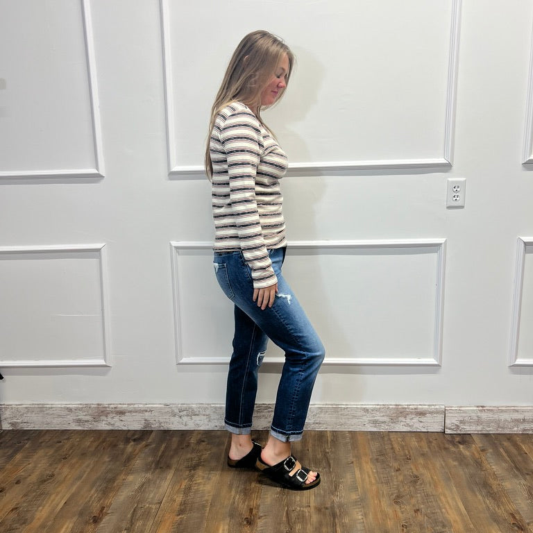 A woman stands in profile wearing a beige and brown striped long-sleeve knit top, blue jeans, and black slide sandals with gold accents, set against a white paneled wall and wood-look floor.