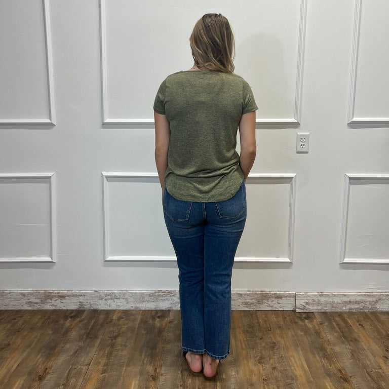 A woman viewed from behind stands on a wood floor against a white paneled wall, wearing an olive green short-sleeve t-shirt and blue jeans.