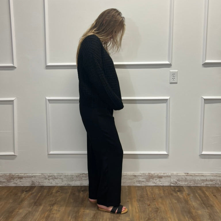 A woman stands in profile wearing a black textured knit long-sleeve top and matching wide-leg pants, standing on a wood floor against a white paneled wall.