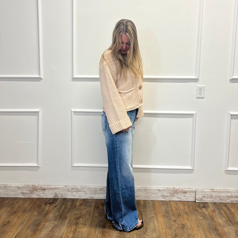 A woman with long blonde hair stands in a room with white paneled walls and a wood floor, wearing a cream ribbed knit cardigan and blue wide-leg jeans.