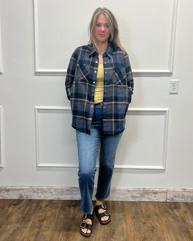 A woman stands in a room with white paneled walls and a wood floor, wearing a blue and gray plaid button-up shirt over a yellow top, blue jeans, and black sandals.