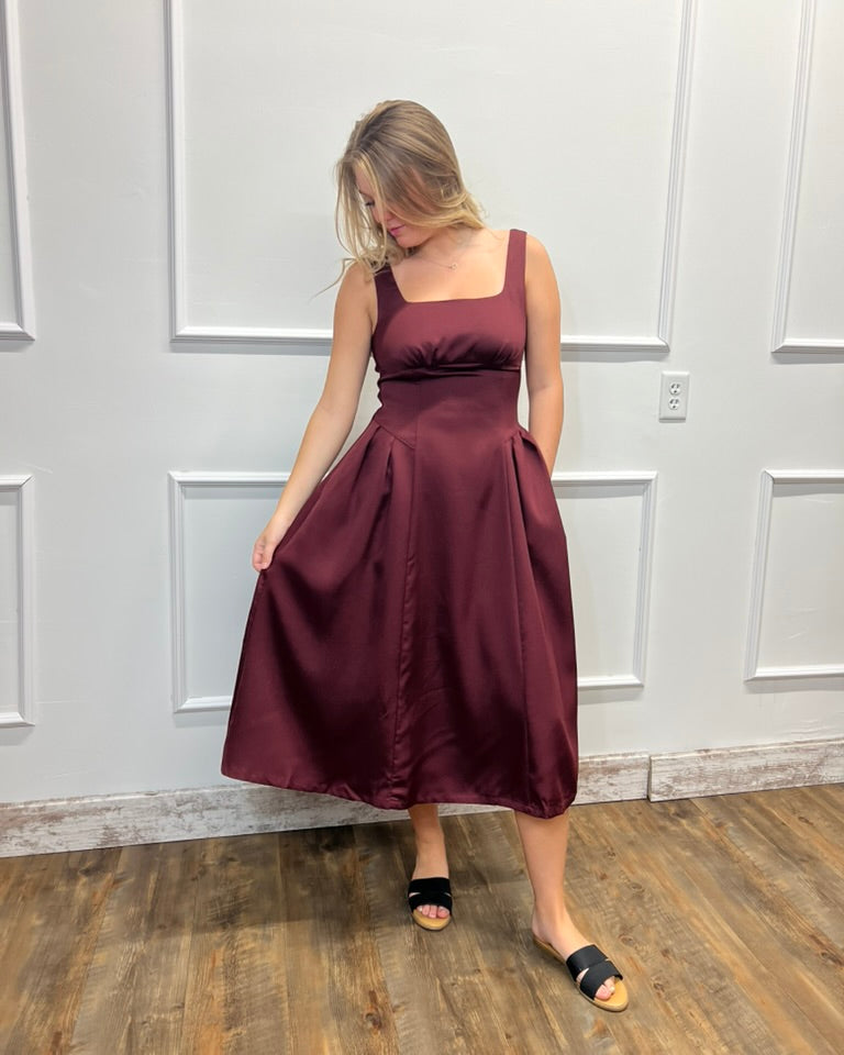 A woman modeling a sleeveless, square-neck midi dress in a deep burgundy satin fabric, standing against a white paneled wall on a wooden floor.