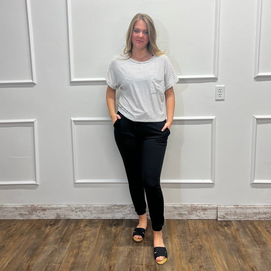 A woman stands against a white paneled wall wearing a white short-sleeve t-shirt with thin black horizontal stripes, black pants, and black slide sandals.