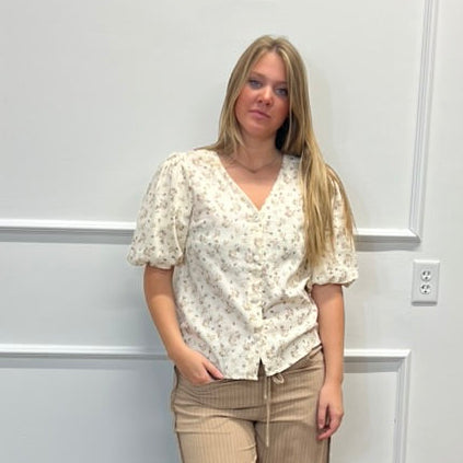 A woman stands in a room with white paneled walls and a wood floor, wearing a cream floral short-sleeve blouse and beige wide-leg pants with black sandals.