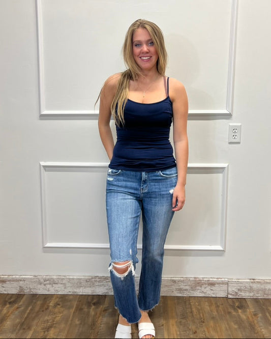 A woman stands indoors against a white paneled wall wearing a dark navy blue ribbed camisole tank top and light-wash distressed blue jeans with ripped knees, paired with white slide sandals.