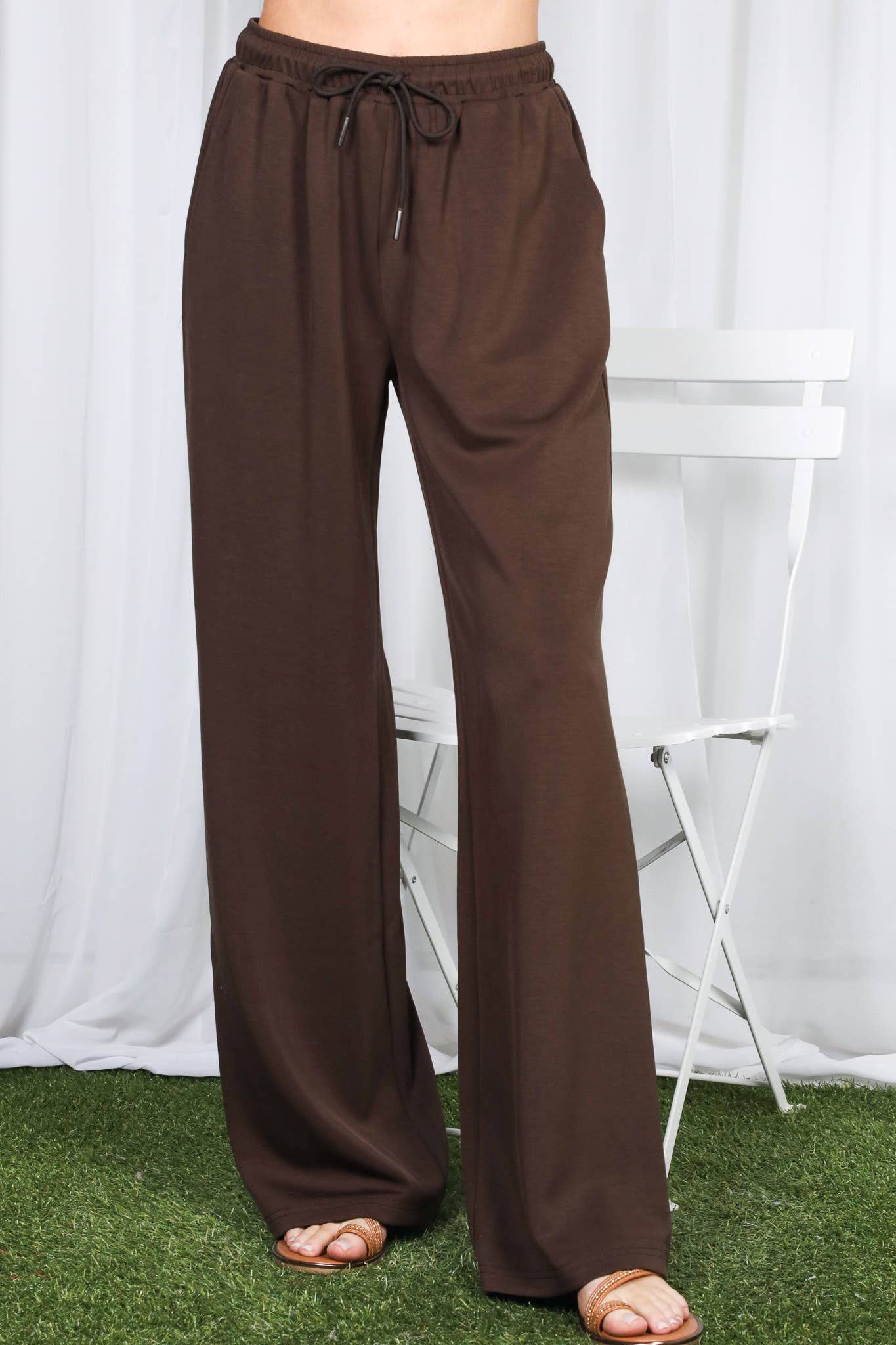 Brown wide-leg drawstring pants shown on a model from the waist down, standing on artificial green grass in front of a white curtain and white folding chair.