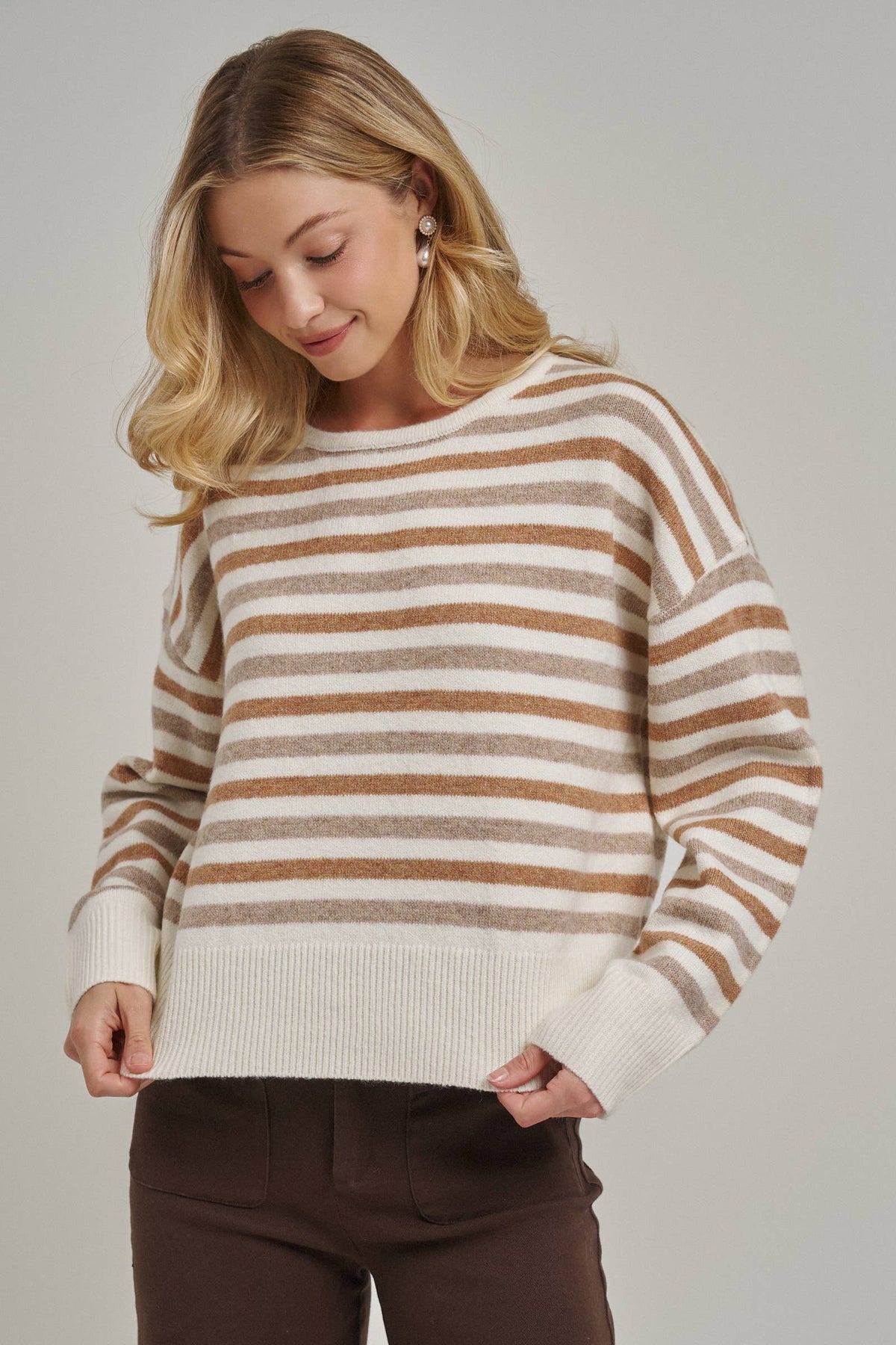 A woman wearing a cream and brown horizontally striped knit sweater with a crew neck and ribbed hem, paired with dark brown pants, photographed against a neutral light gray studio background.