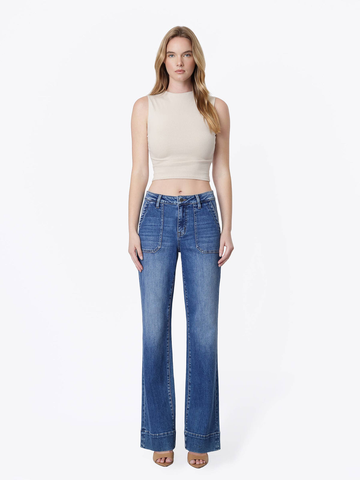 A woman modeling a pair of high-waisted, wide-leg blue jeans with a beige sleeveless crop top, standing against a plain white studio background.