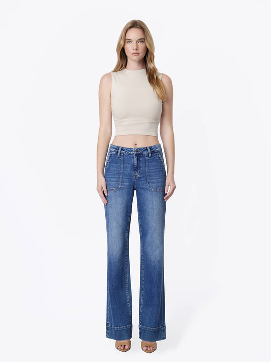 A woman modeling a pair of high-waisted, wide-leg blue jeans with a beige sleeveless crop top, standing against a plain white studio background.