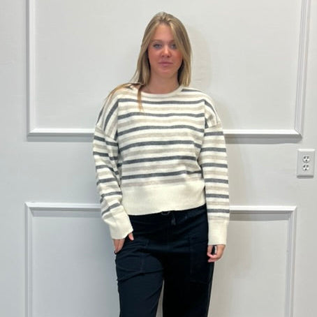 A woman stands in a room with white paneled walls and a wood floor, wearing a cream and gray striped knit sweater, black pants, and black slide sandals.