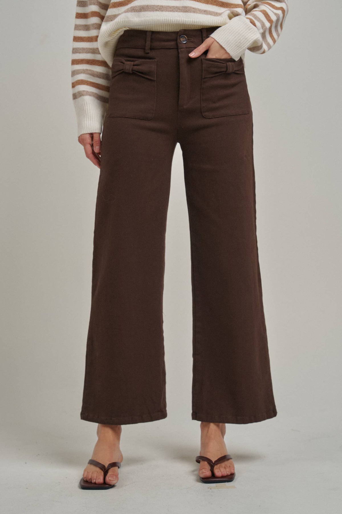 A model is wearing high-waisted, wide-leg brown pants paired with a cream and brown striped sweater and dark brown thong sandals, photographed against a neutral light gray studio background.
