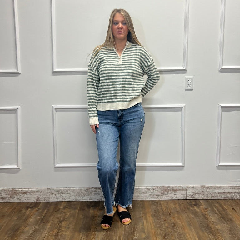 A woman stands in a room with white paneled walls and a wood floor, wearing a green and white striped half-zip sweater, blue jeans, and black slide sandals.