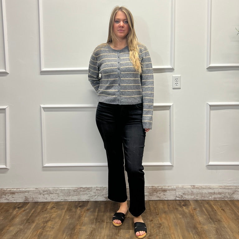 A woman stands in a room with white paneled walls and wood floors, wearing a gray and white striped knit sweater, black pants, and black slide sandals.