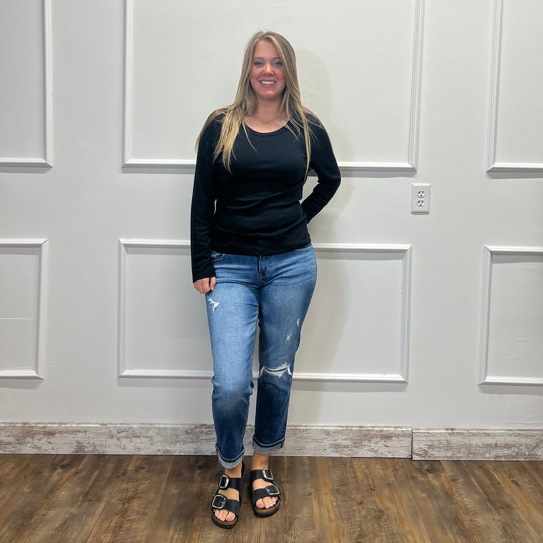 A woman stands in front of a white paneled wall wearing a black long-sleeve top, blue distressed jeans, and black sandals.