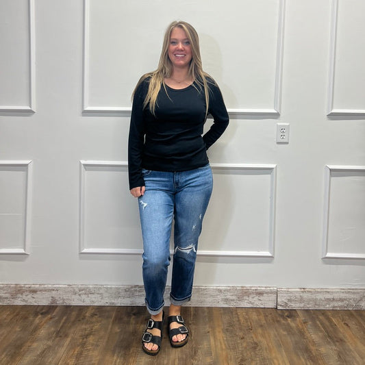 A woman stands in front of a white paneled wall wearing a black long-sleeve top, blue distressed jeans, and black sandals.