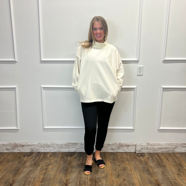 A woman modeling a cream mock-neck sweatshirt with side cutouts, paired with black pants and black slide sandals, standing in front of a white paneled wall on a wood-look floor.