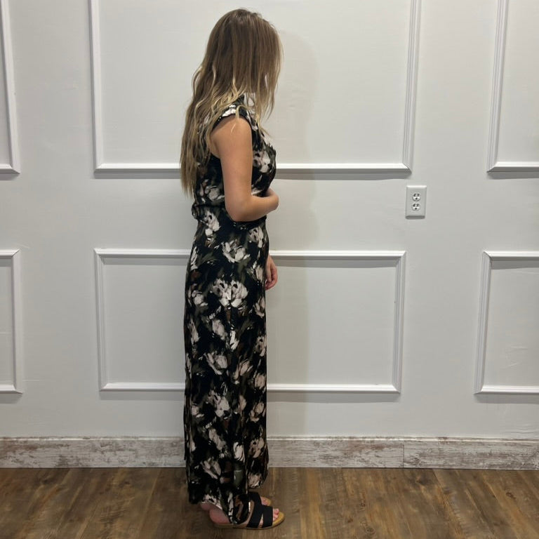 A woman stands in profile wearing a sleeveless black maxi dress with a white floral pattern, standing on a wood floor against a white paneled wall.
