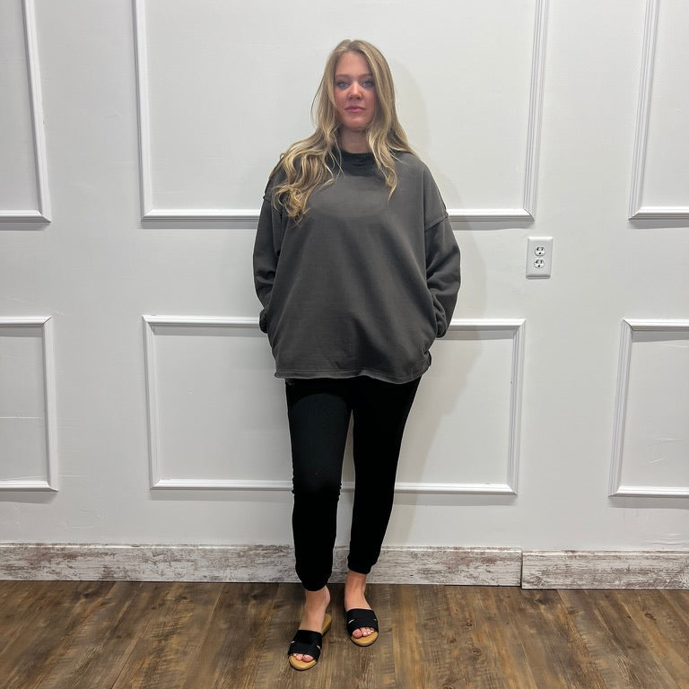 A woman stands in front of a white paneled wall wearing an oversized dark gray sweatshirt, black pants, and black slide sandals.