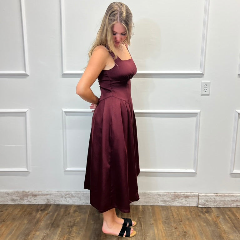A woman modeling a sleeveless, high-waisted, midi-length A-line dress in a deep burgundy or maroon color, standing against a white paneled wall with a wood floor.