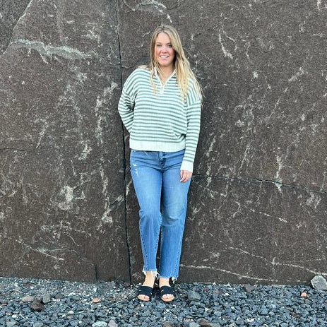 A woman stands against a large dark stone wall outdoors, wearing a light green and white striped long-sleeve top, blue jeans, and black sandals, with gravel in the foreground and trees behind her.