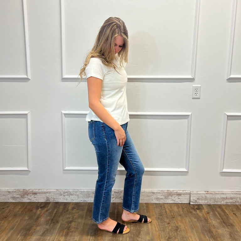 A woman stands in profile wearing a white short-sleeve t-shirt, blue straight-leg jeans, and black slide sandals, set against a white paneled wall and a wood-look floor.